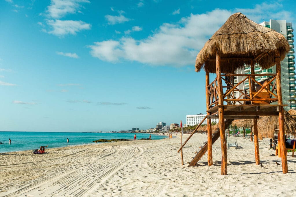 Sandy beach scene with thatched lifeguard tower, turquoise sea, people walking, and distant skyscrapers against a clear blue sky.