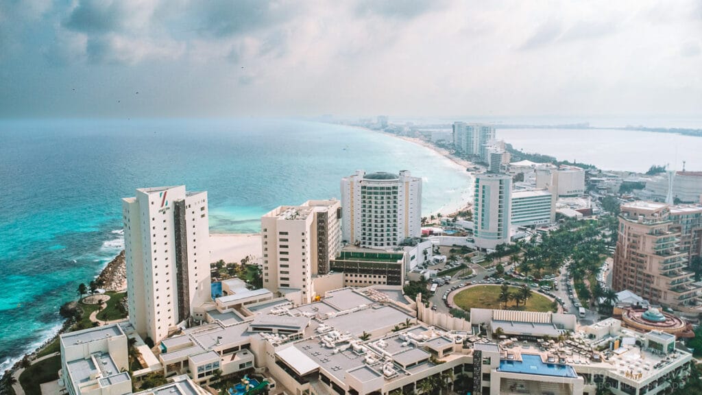 Aerial view of Cancún's hotel zone with high-rise buildings, turquoise ocean, and white sandy beach under a partially cloudy sky.