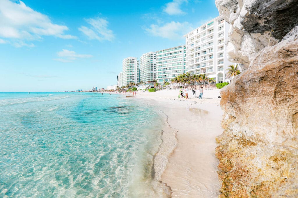 Clear turquoise waters and sandy beach in Cancún, Mexico; high-rise buildings and people strolling along the shore under a bright blue sky.