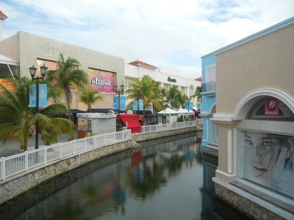 Centro comercial al aire libre con un canal que lo atraviesa, rodeado de palmeras y escaparates de colores brillantes.