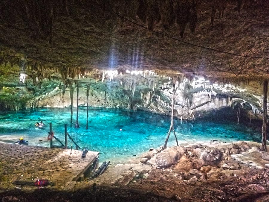 People swimming in a stunning, turquoise cenote cave with stalactites, surrounded by natural rock formations and illuminated by sunlight filtering from above.