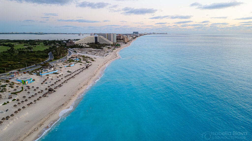 Aerial view of Cancun's coastline with turquoise water, hotel resorts, sandy beaches, and palapas. The landscape stretches into the distance under a cloudy sky.