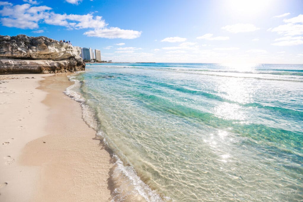 A serene beach scene with clear turquoise water, rocky cliffs, and distant buildings under a bright blue sky with scattered clouds.