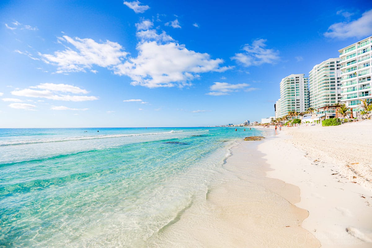 Clear blue skies, turquoise ocean waves, sandy beach, and modern high-rise buildings line the shore, with a few people strolling along the coast.