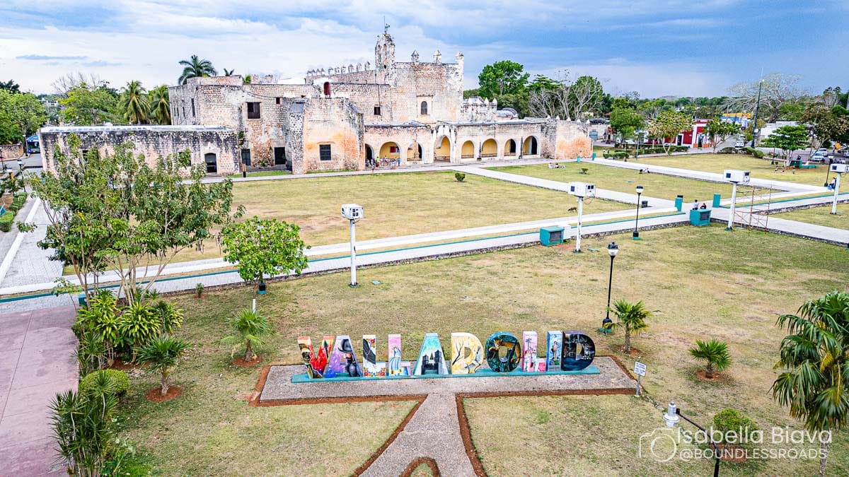 A historic stone building, San Bernardino de Siena Convent, stands in a spacious park with colorful "Valladolid" letters, trees, and pathways around it.