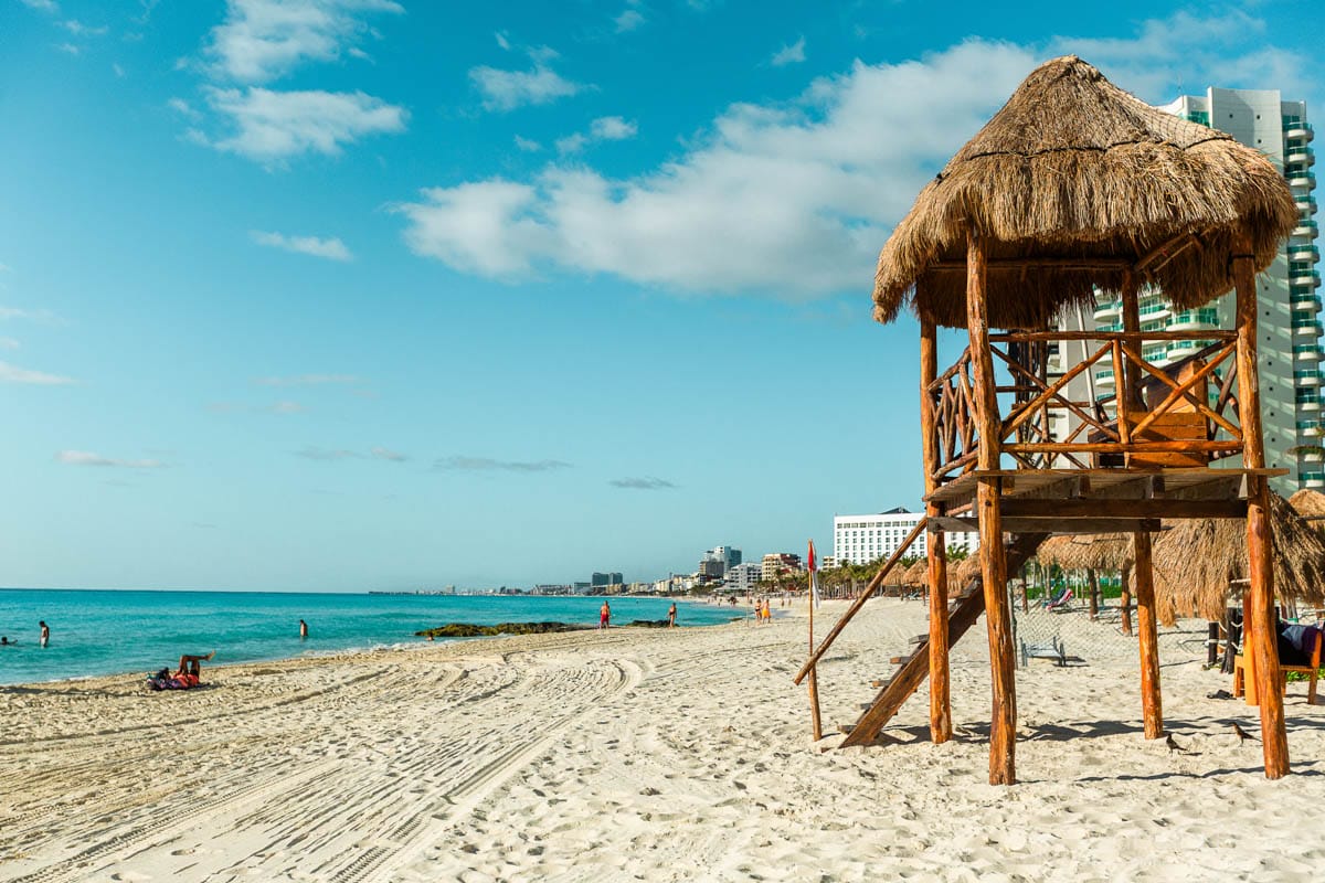 A sandy beach with a wooden lifeguard hut, bright blue ocean, several buildings, and a few people enjoying the sunny day.