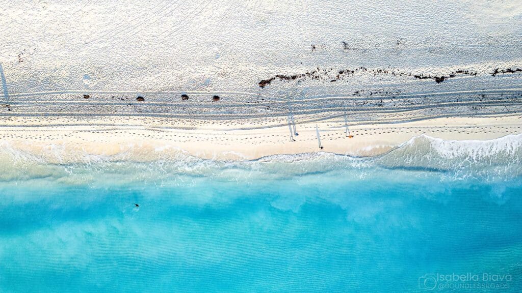 Aerial view of a pristine beach with turquoise water, white sand, and parallel footprints lining the shore in a tranquil setting.