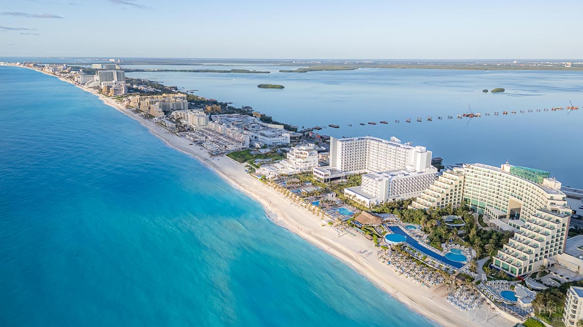 Aerial view of Cancún's hotel zone featuring turquoise waters, sandy beach, and luxurious resorts against a clear blue sky with distant land formations.