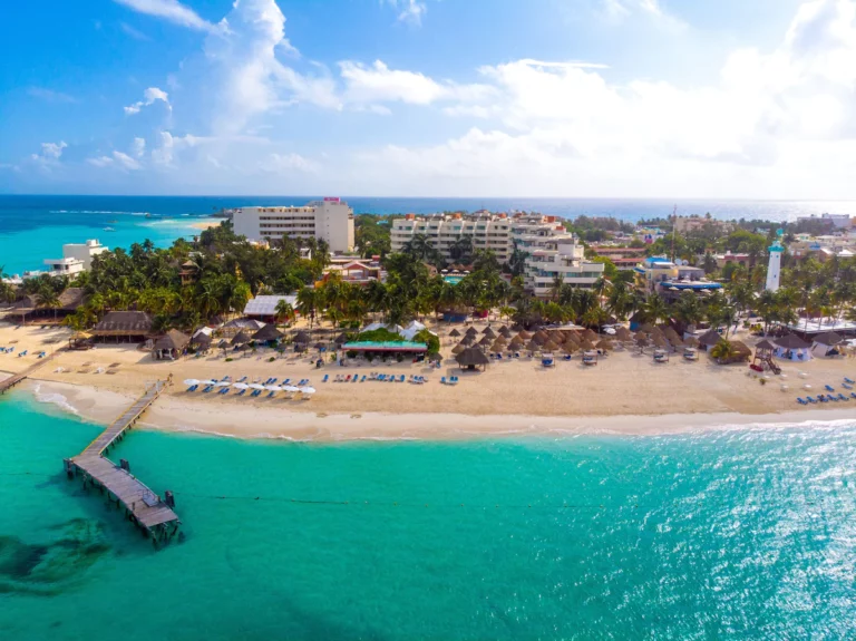 Aerial view of a tropical beach with turquoise waters, lined with lounge chairs and straw umbrellas. A wooden pier extends into the ocean. In the background, there are several luxury hotels and palm trees on Isla Mujeres, under a partly cloudy sky.