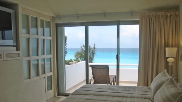 A bedroom with beige walls and linens features a sliding glass door that opens to a balcony with a lounge chair. The balcony overlooks the turquoise ocean and a blue sky with a few clouds. This Airbnb in Cancun also has a wall-mounted TV on the left side for your entertainment.