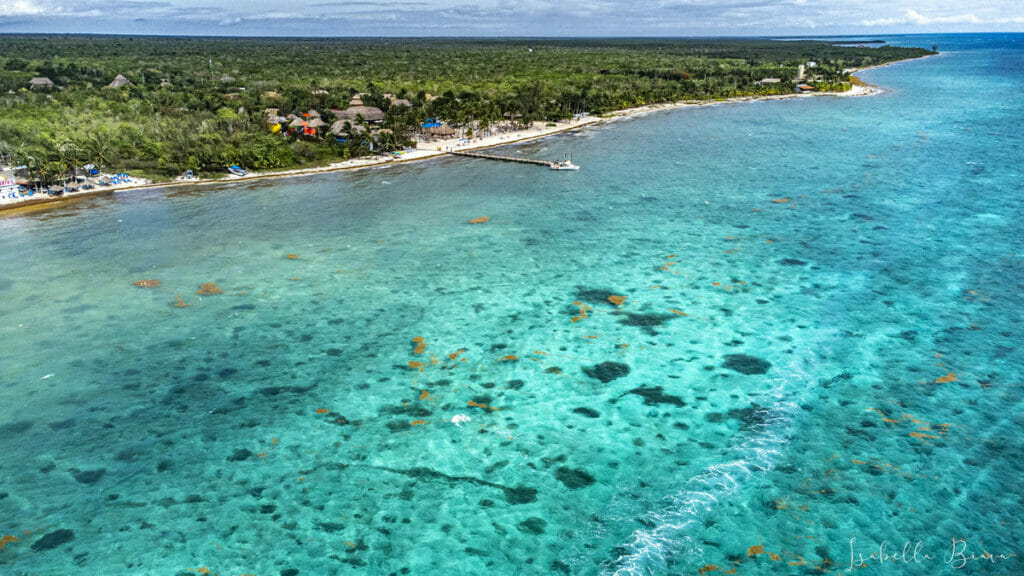 Aerial view of a tropical coastline with turquoise waters, a pier extending into the ocean, and dense greenery along the shore. Small buildings and boats are visible near the water.