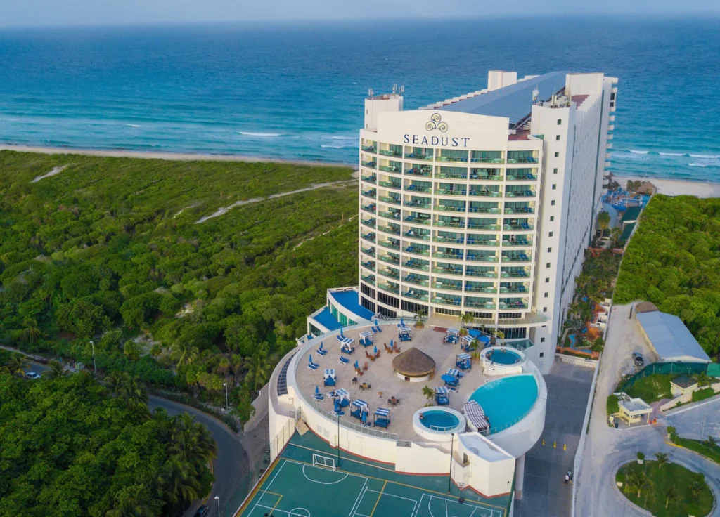 Seadust Cancun Family Resort - tall hotel with circular shaped lounging area in front and three pools, beach is behind the hotel 