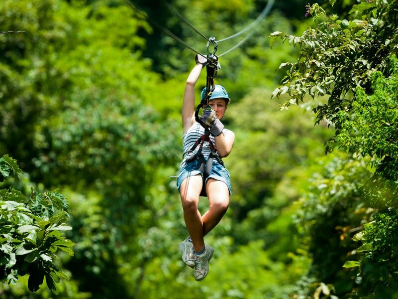A person wearing a helmet and harness glides on a zipline through a lush, green forest, reminiscent of the thrill you find on ATV tours in Cancun.