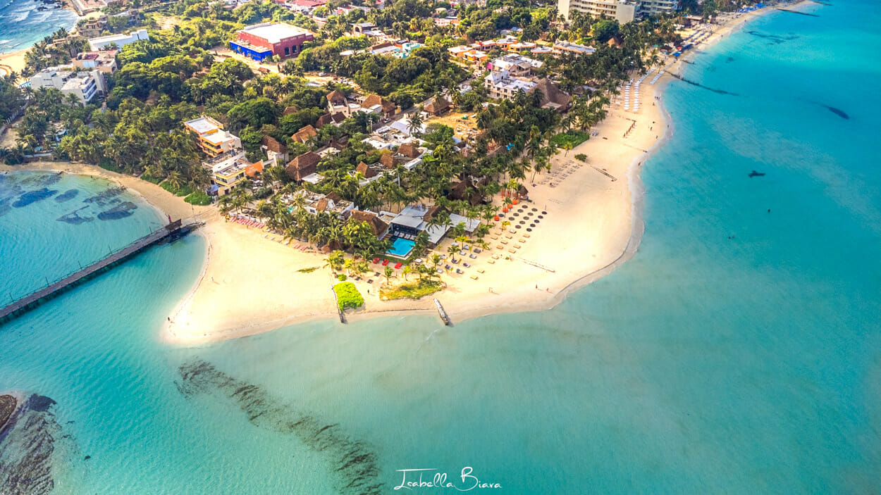 Aerial view of a coastal area with a sandy beach, turquoise waters ideal for Cancun Snorkeling Tours, and numerous beach chairs and umbrellas. Lush green trees and buildings are visible inland, along with a small pier extending into the water.
