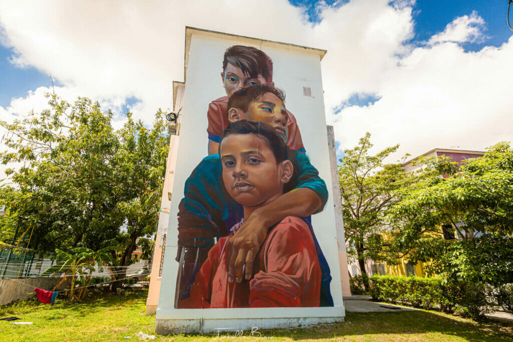 A large mural depicts a woman hugging two children. They are painted on the side of a tall, rectangular building. The background features a bright blue sky with clouds and greenery around the building.