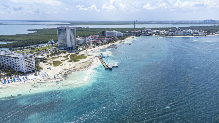Aerial view of Cancun's hotel zone with white sandy beaches, numerous high-rise hotels, a pier with docked boats, and a mix of turquoise and deep blue ocean waters. Nearby green landmasses are visible.
