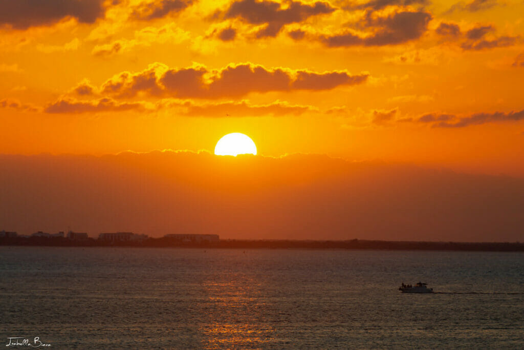 A vibrant sunset over the ocean in Cancun in winter, with the sun partially hidden behind clouds. A small boat is visible on the water, leaving a subtle wake. The sky is a gradient of orange and yellow hues.