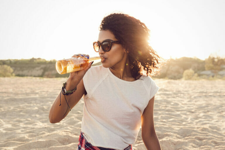 A woman in white top drinking on the beach.