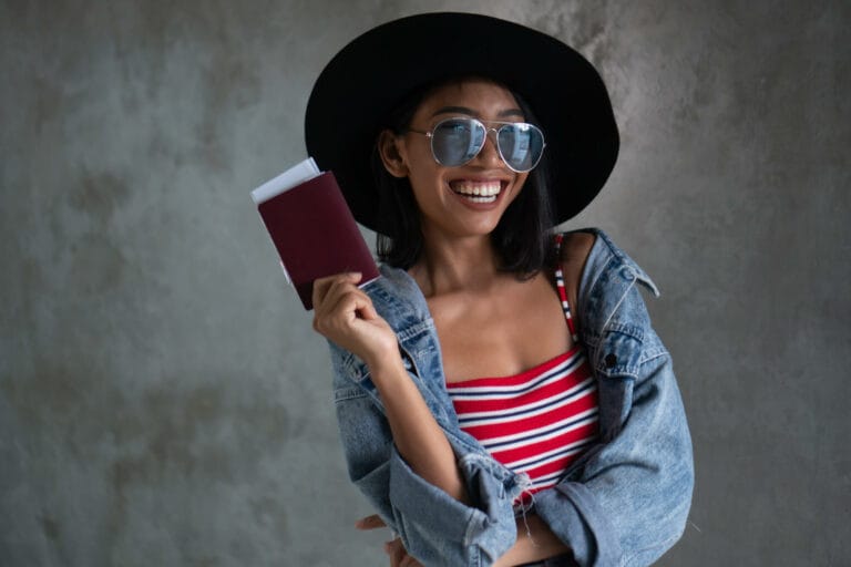A woman smiles broadly while holding a passport, almost as if she's ready to answer, "Do you need a passport to go to Cancun?" She wears a black wide-brimmed hat, sunglasses, a striped top, and a denim jacket hanging off her shoulders. The background is a plain gray wall.