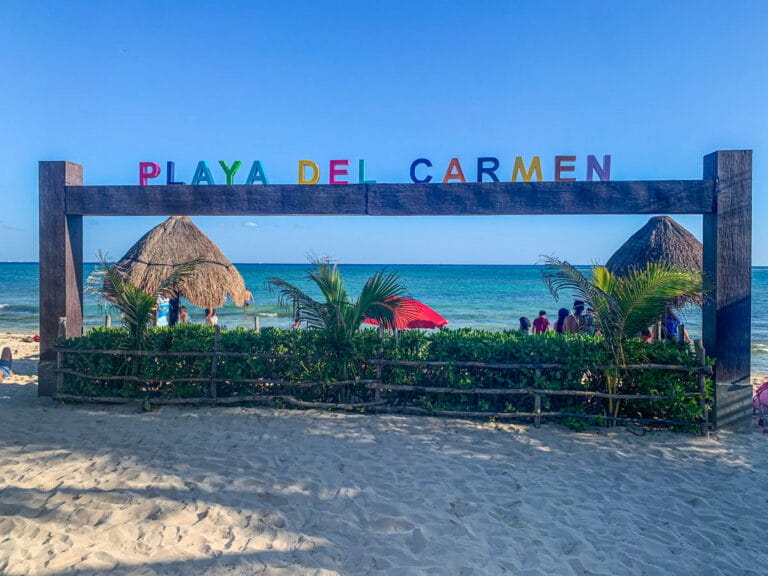 Colorful "Playa Del Carmen" sign on a wooden frame at a sandy beach with palapa huts, people, palm trees, and the clear blue sea in the background under a bright blue sky—perfect for those seeking adventurous things to do in Cancun.