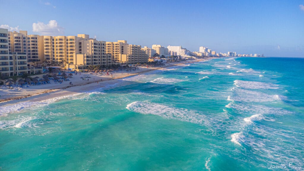 Aerial view of a coastal city with numerous beachfront buildings and resorts lining the shore. The turquoise ocean waves crash onto the sandy beach under a clear blue sky.