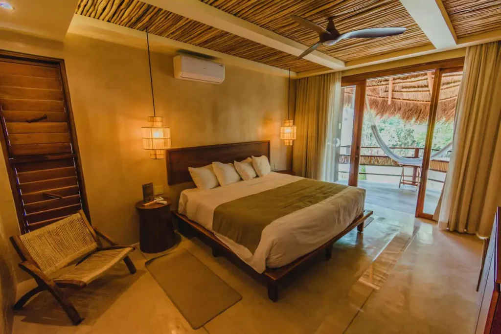 A cozy bedroom with a large bed, wooden ceiling, ceiling fan, two nightstands with lamps, a chair, and an open door leading to a balcony with a hammock. Neutral tones create a warm ambiance reminiscent of Bacalar in Mexico – the perfect retreat recommended by any travel guide.
