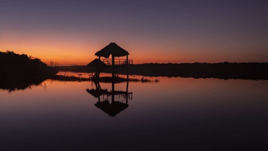 Silhouetted huts on stilts over a calm lake at sunset in Bacalar, Mexico. The sky is a gradient from deep purple to orange, reflecting on the water, creating a tranquil and serene scene perfect for your 2024 travel guide.