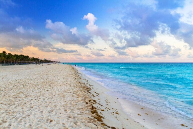 A picturesque beach with turquoise waters, soft white sand, and a partly cloudy sky at sunset.