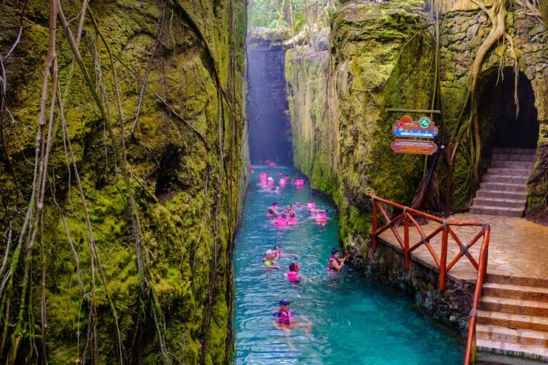 People swim through a narrow, lush canyon with turquoise water, surrounded by mossy walls. Wooden railing and stairs lead to the entrance.