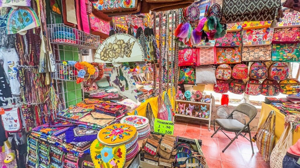 A vibrant market stall filled with colorful textiles, bags, and handcrafted items, showcasing intricate patterns and traditional designs. Two chairs are visible.
