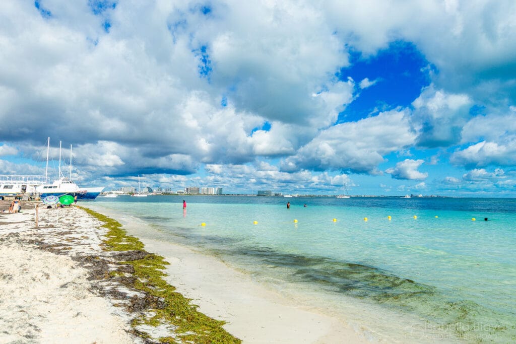 Shallow, crystal-clear beach water under a partly cloudy blue sky with a sandbar stretching into the sea.