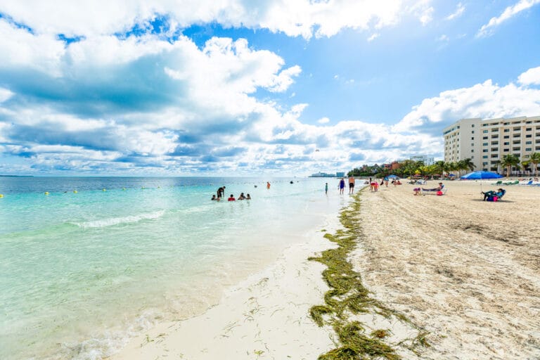 Shallow, crystal-clear beach water under a partly cloudy blue sky with a sandbar stretching into the sea and people swimming