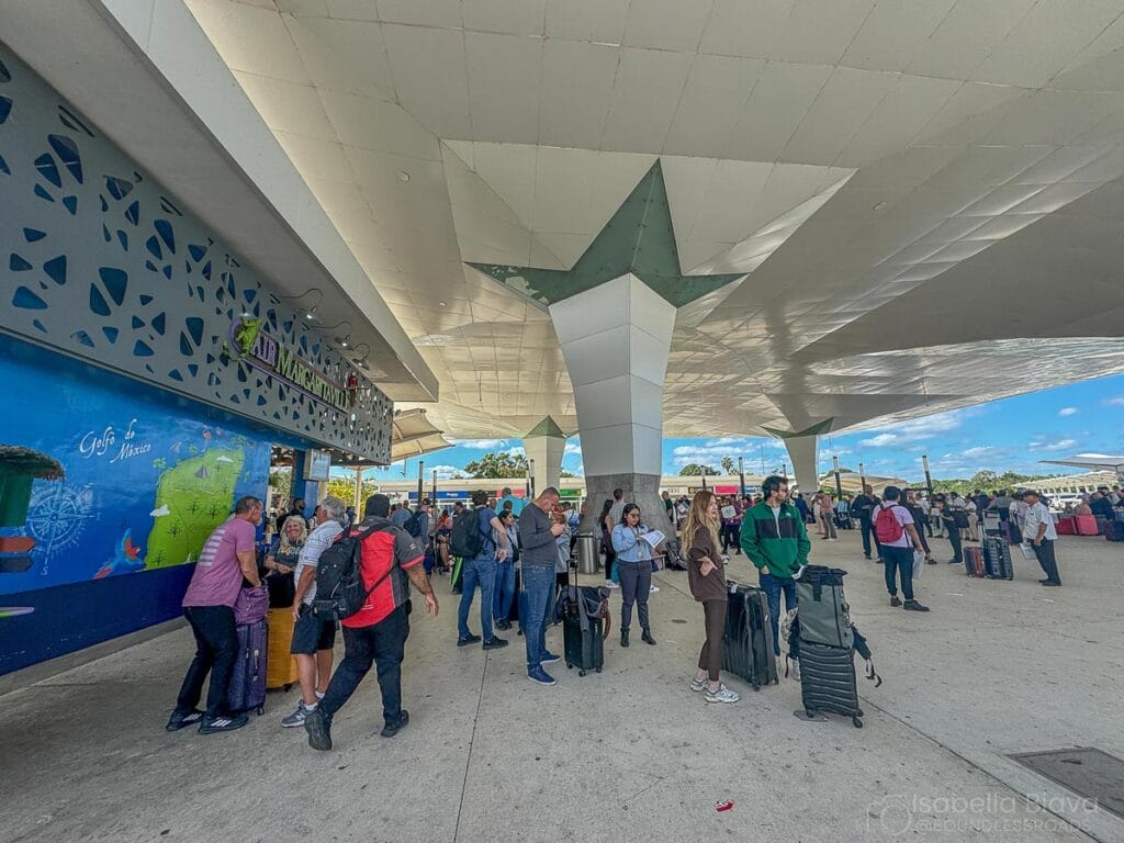 People with luggage gather outside a modern airport terminal, featuring a star-shaped roof structure and colorful murals, under a clear blue sky.