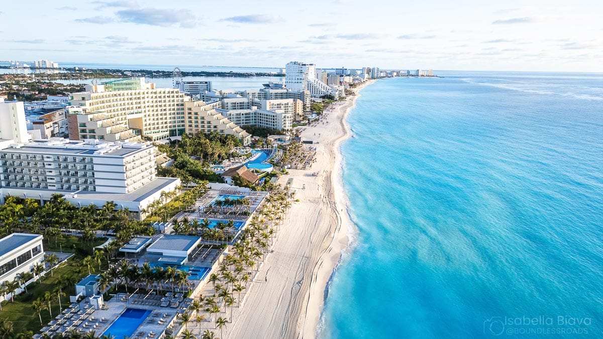 Aerial view of Cancun's beachfront hotels, clear blue ocean on the right, with palm trees and people enjoying the sandy shore.