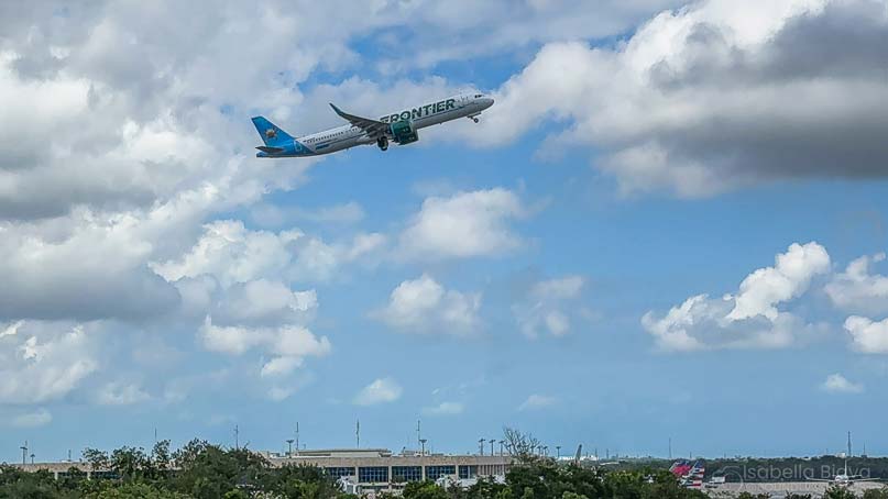 A Frontier Airlines plane ascends against a partly cloudy sky above a landscape with buildings and trees, indicating an airport environment.