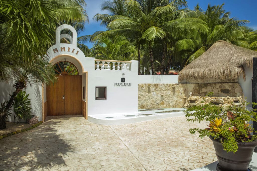 Tropical resort entrance with palm trees and thatched roof.